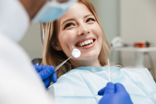 Slika mlade nasmejane ženske, ki sedi na zobozdravstvenem stolu. Obravnava jo prijazen zobozdravnik.
